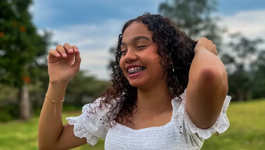 Jovem de blusa branca sorri com a mão no cabelo em um gramado.