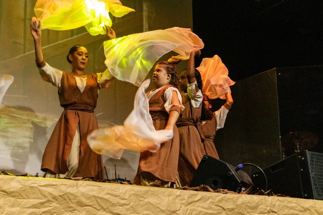 Atrizes de figurino marrom agitam tecidos amarelos no palco iluminado.