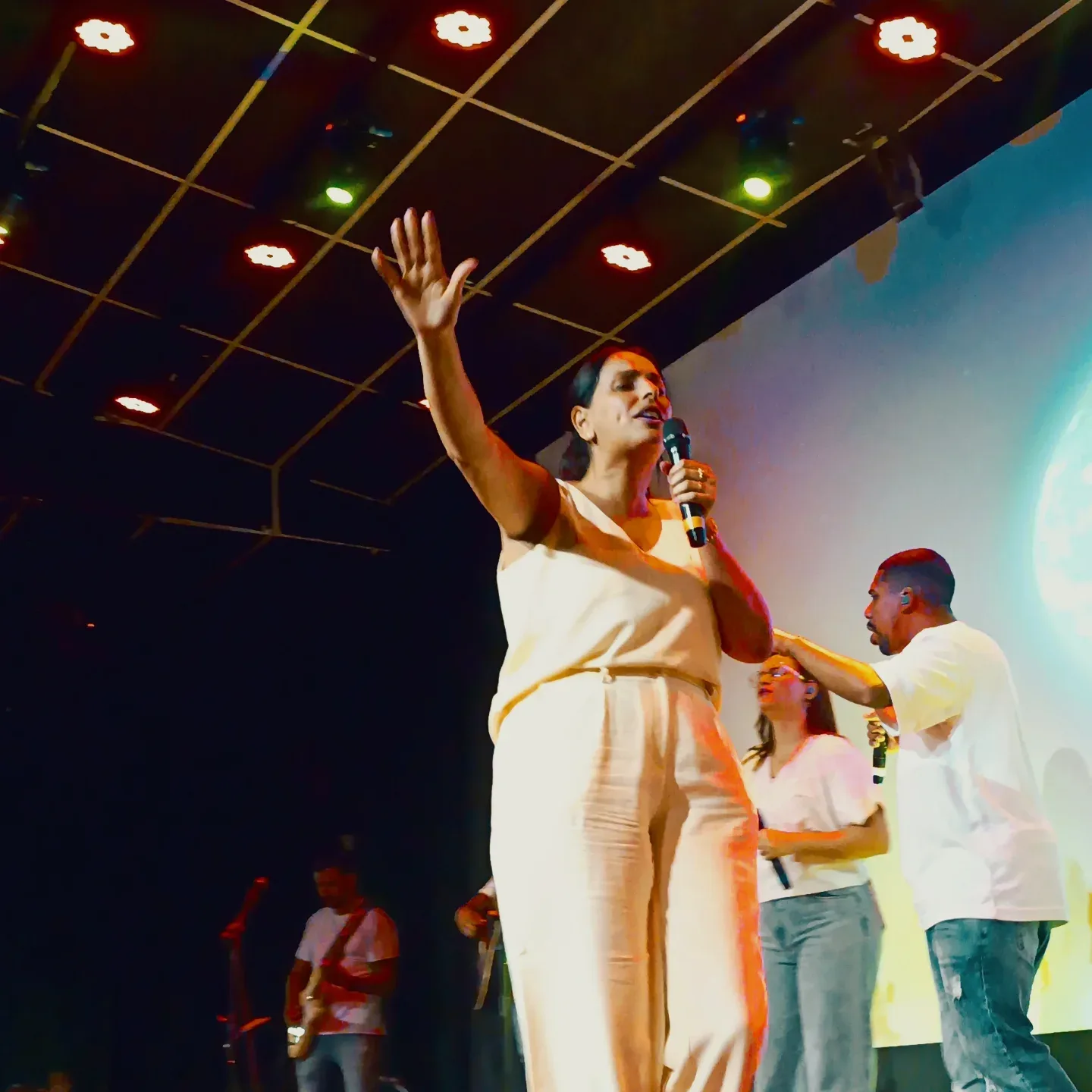 Mulher de roupa clara fala ao microfone com o braço erguido em um palco com luzes coloridas.
