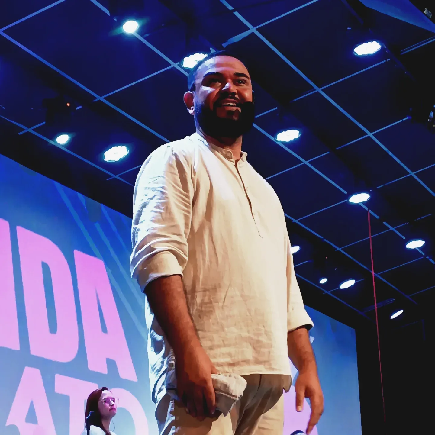 Homem de camisa branca no palco, com telão azul e rosa ao fundo.