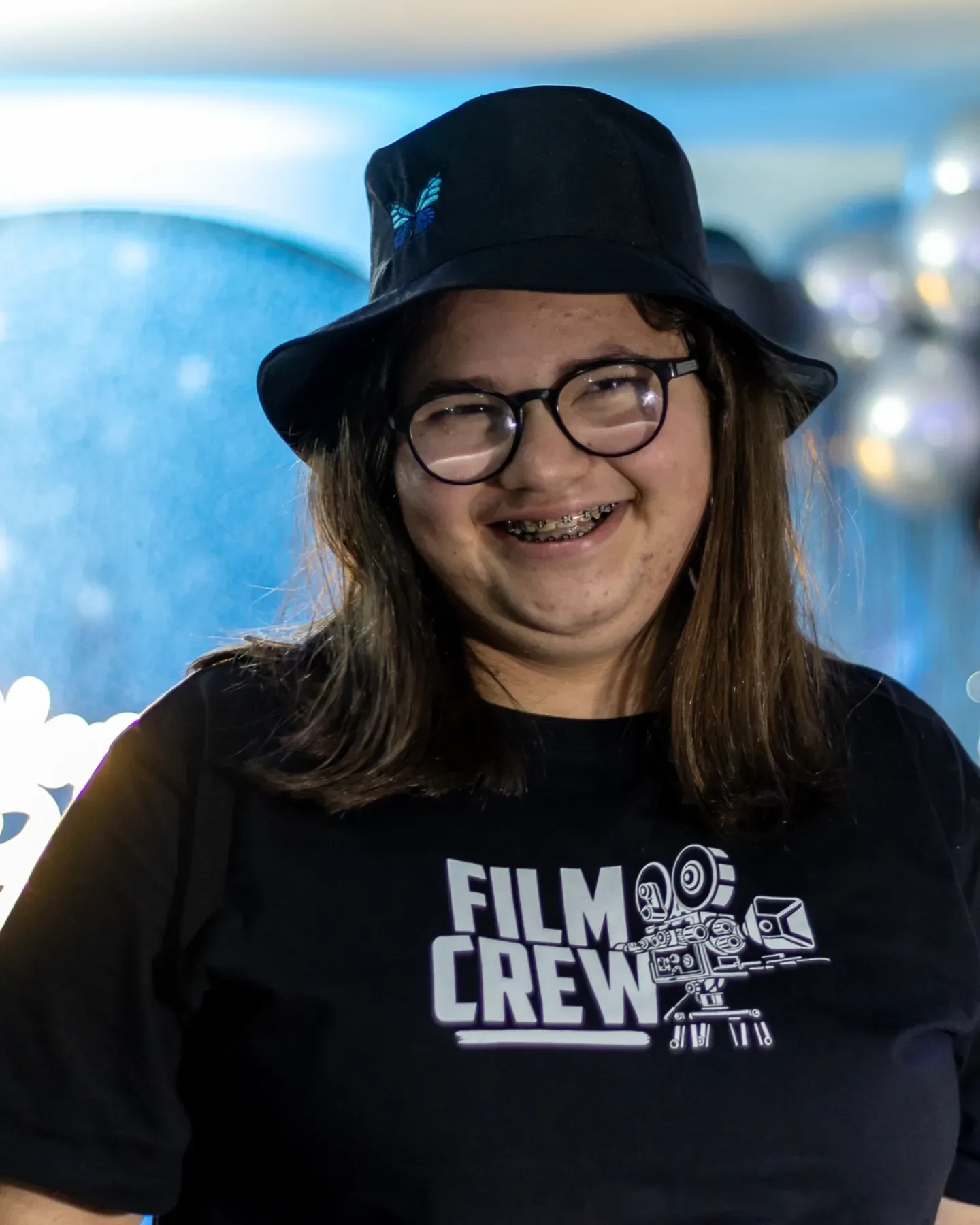 Retrato de Jhenifer sorrindo, de chapéu preto, óculos e camiseta preta, diante de um fundo azul.
