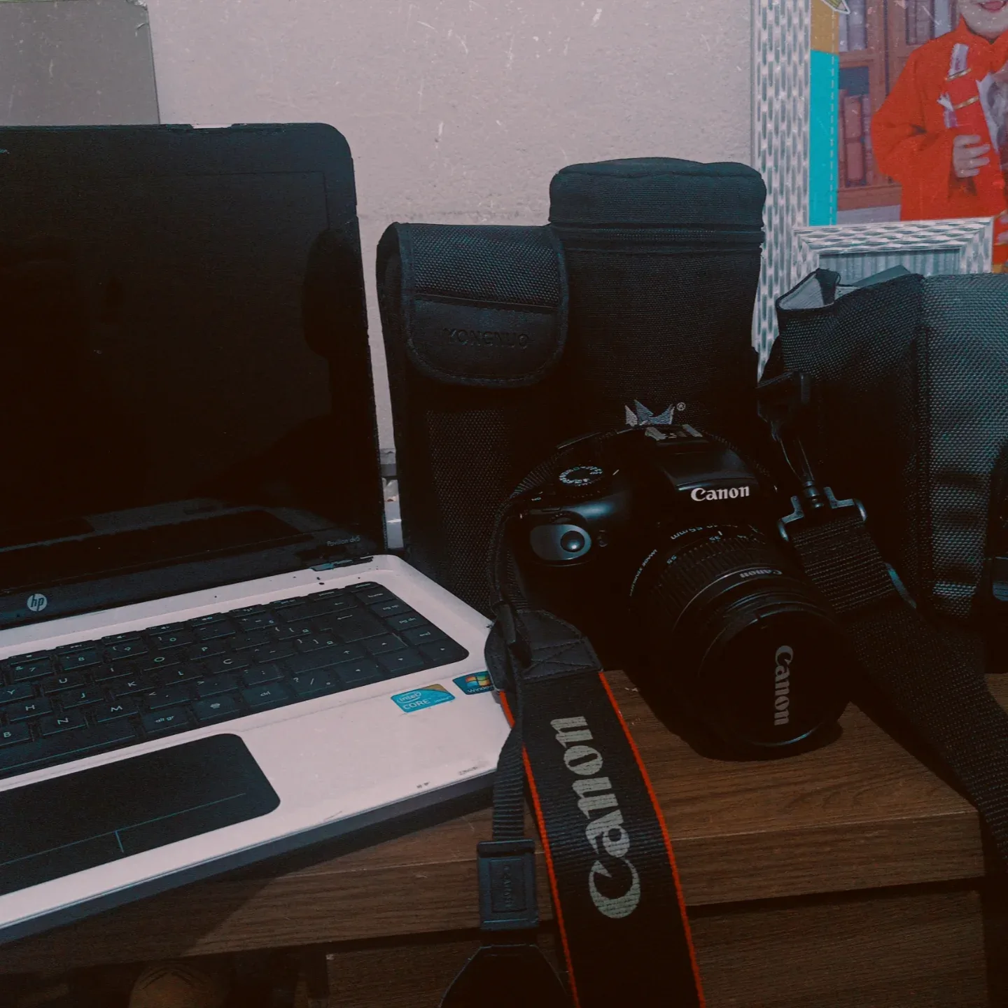 Notebook aberto ao lado de uma câmera Canon e bolsas de equipamento sobre a mesa.