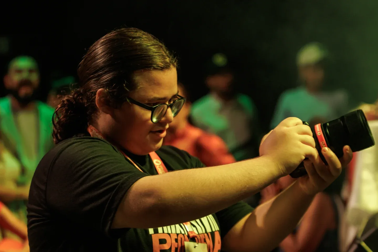 Videomaker de óculos enquadra uma cena com a câmera durante um evento, com luzes verdes ao fundo.