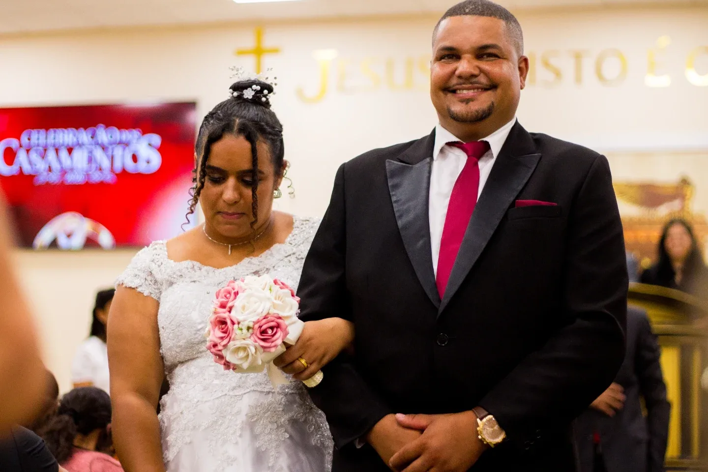 Noiva com buquê e noivo de terno sorrindo lado a lado durante a cerimônia de casamento.