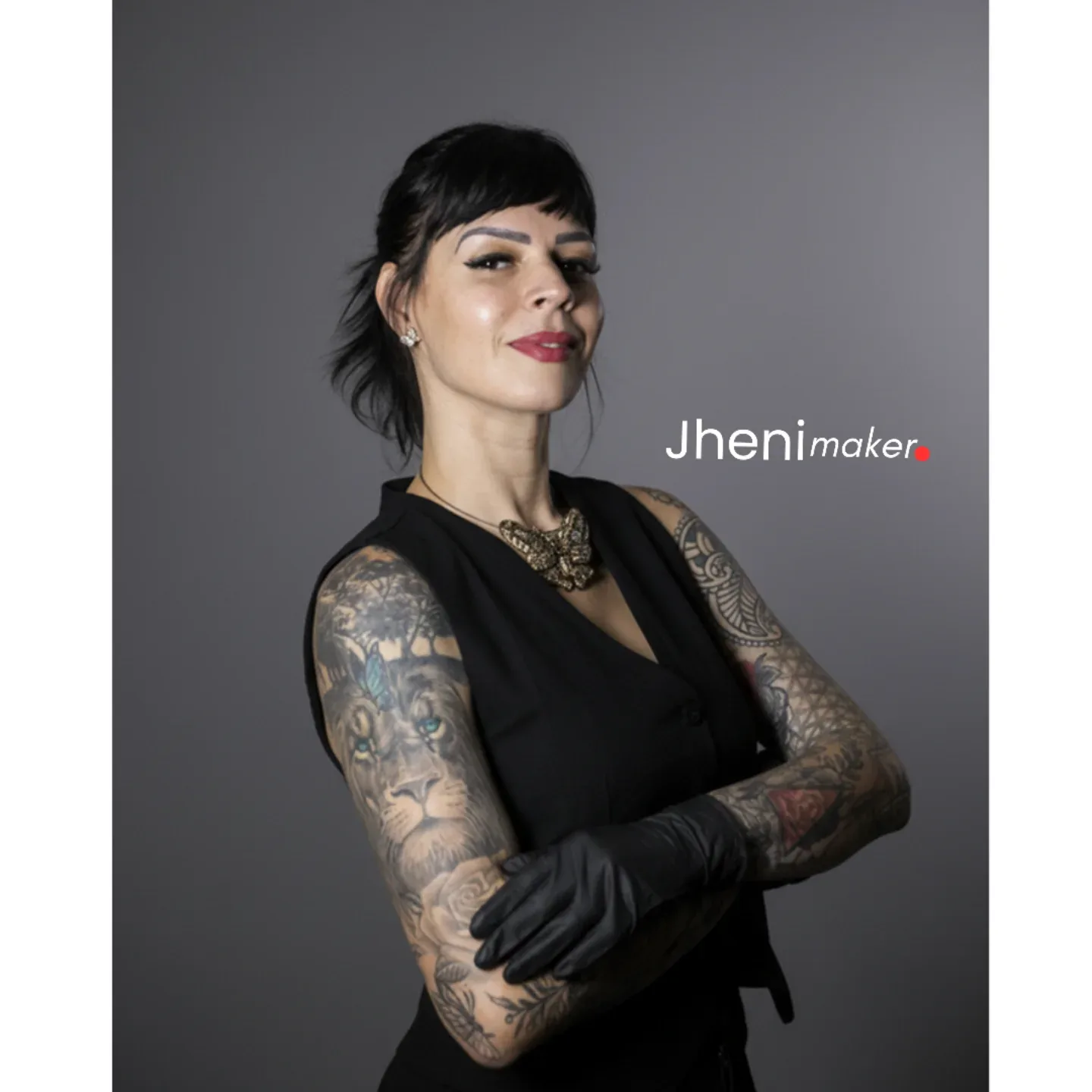 Retrato de uma mulher de cabelo preto curto, braços tatuados e luvas pretas, diante de um fundo cinza.